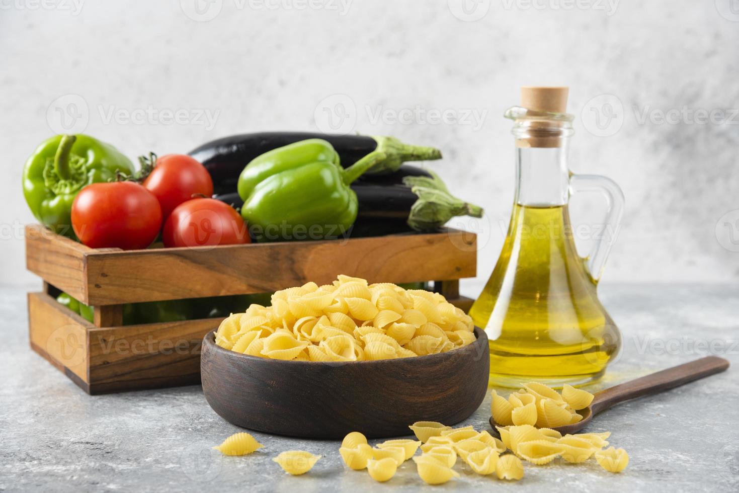 pasta cruda en forma de concha con una botella de vidrio de aceite y una caja de madera llena de verduras frescas foto