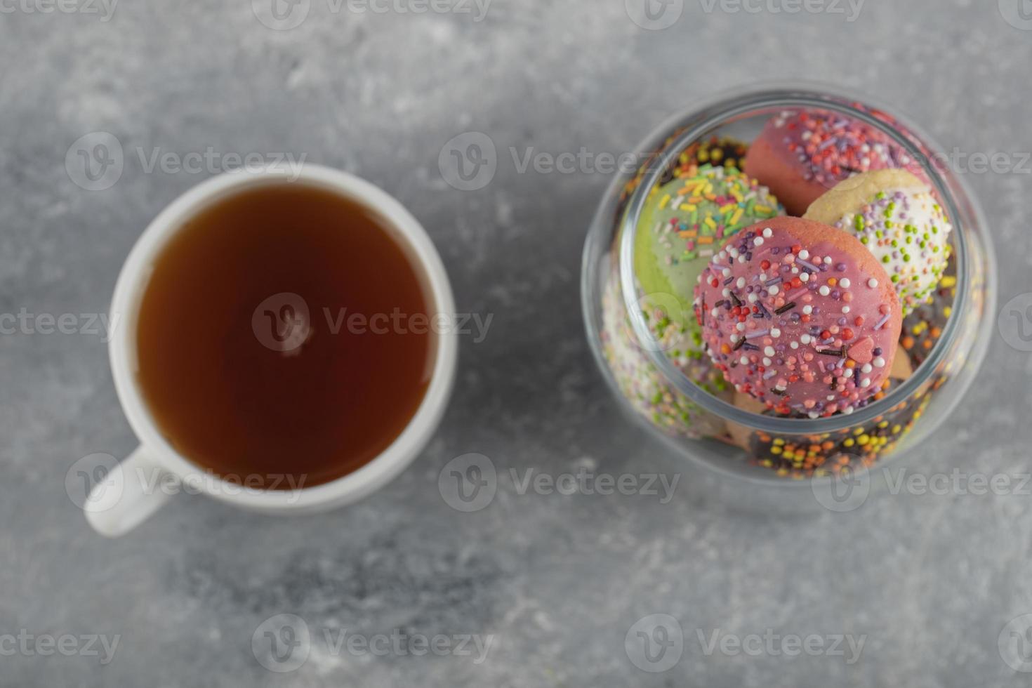 un frasco de vidrio lleno de pequeñas rosquillas de colores con una taza de té caliente foto