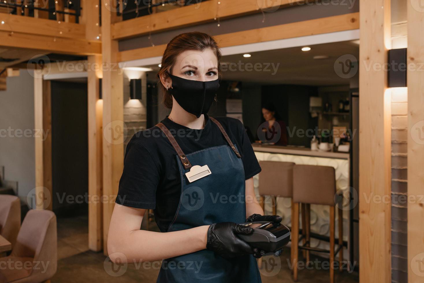Una camarera que usa un delantal, una mascarilla médica negra y guantes médicos desechables sostiene un terminal de pago inalámbrico en un restaurante. foto