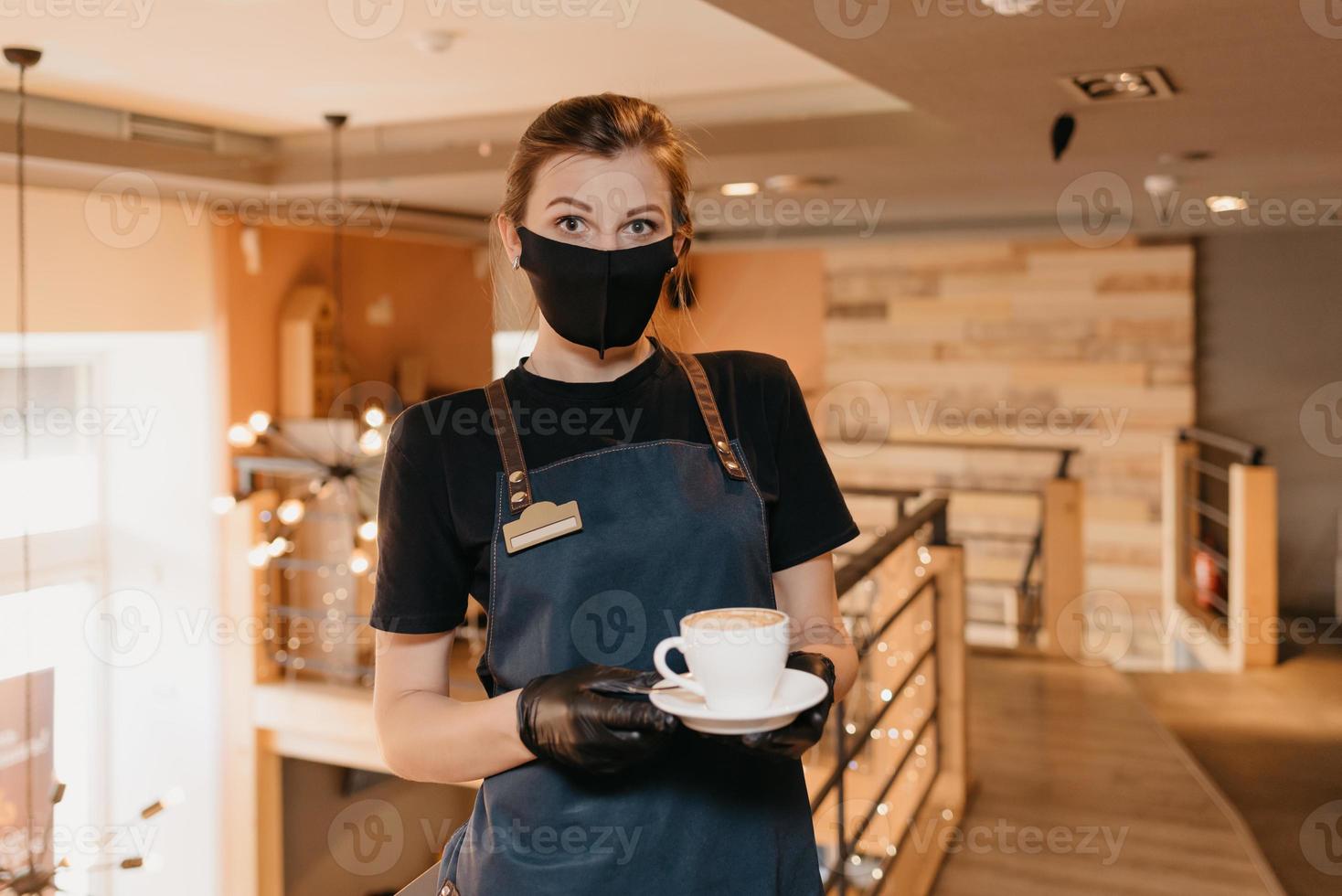 A waitress who wears a black face mask and disposable gloves serves clients in a cafe photo