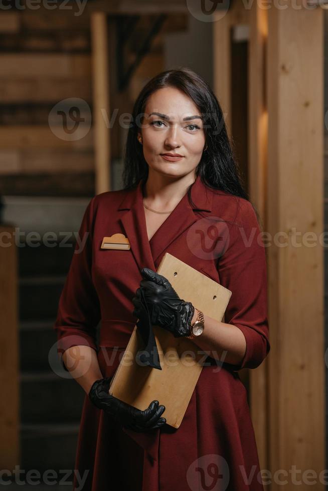 A manager in black disposable medical gloves wears a medical face mask holding a wooden menu in a restaurant photo