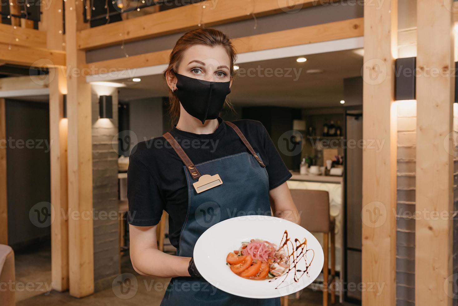 A stylish waitress who wears a black face mask and disposable gloves is holding a mint and dolendwitz salad in a restaurant photo