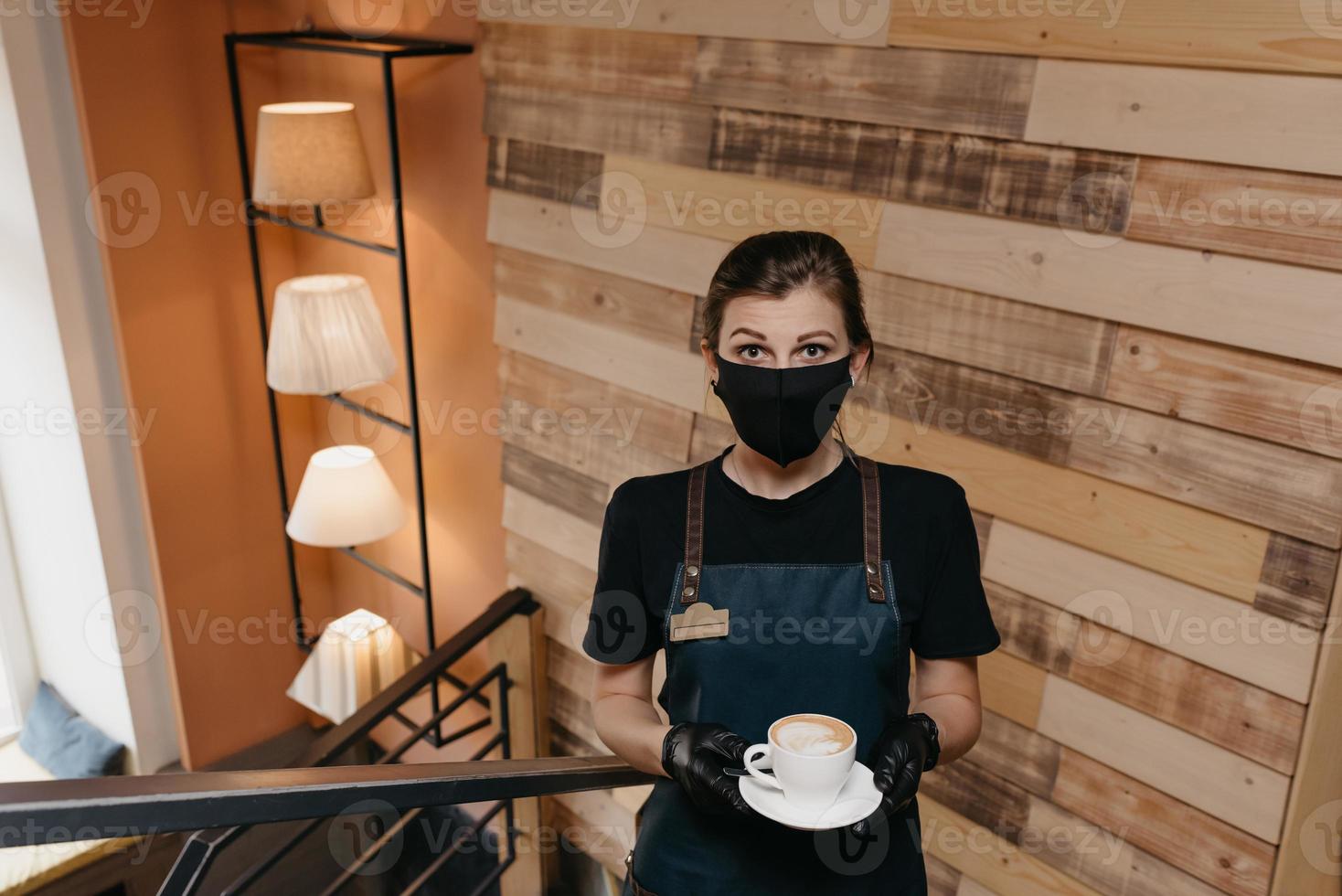 A waitress who wears a black face mask and disposable gloves serves clients in a cafe photo