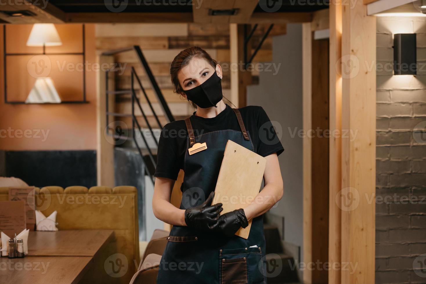 Una camarera con guantes médicos desechables negros lleva una mascarilla médica sosteniendo un menú de madera en un restaurante foto