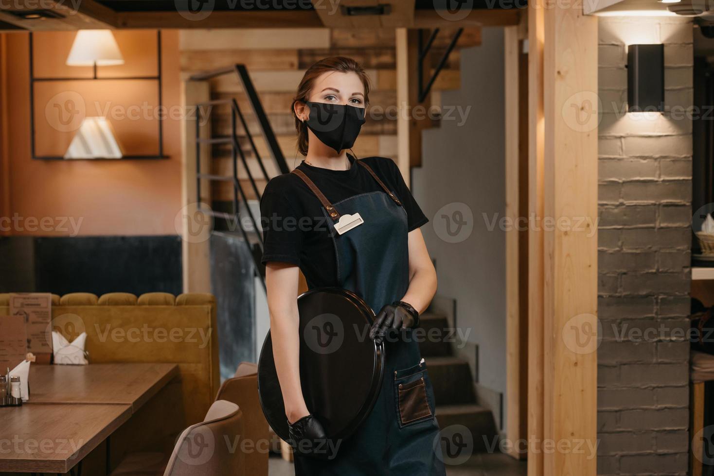 Una camarera lleva una mascarilla médica y guantes médicos desechables negros posando con una bandeja de plástico en un restaurante. foto