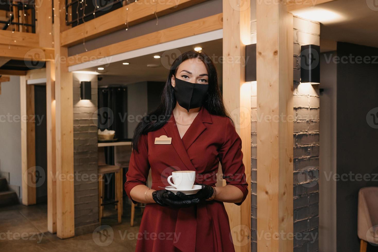 A female restaurant manager wears a black face mask and disposable gloves holding a cup of coffee in a restaurant photo