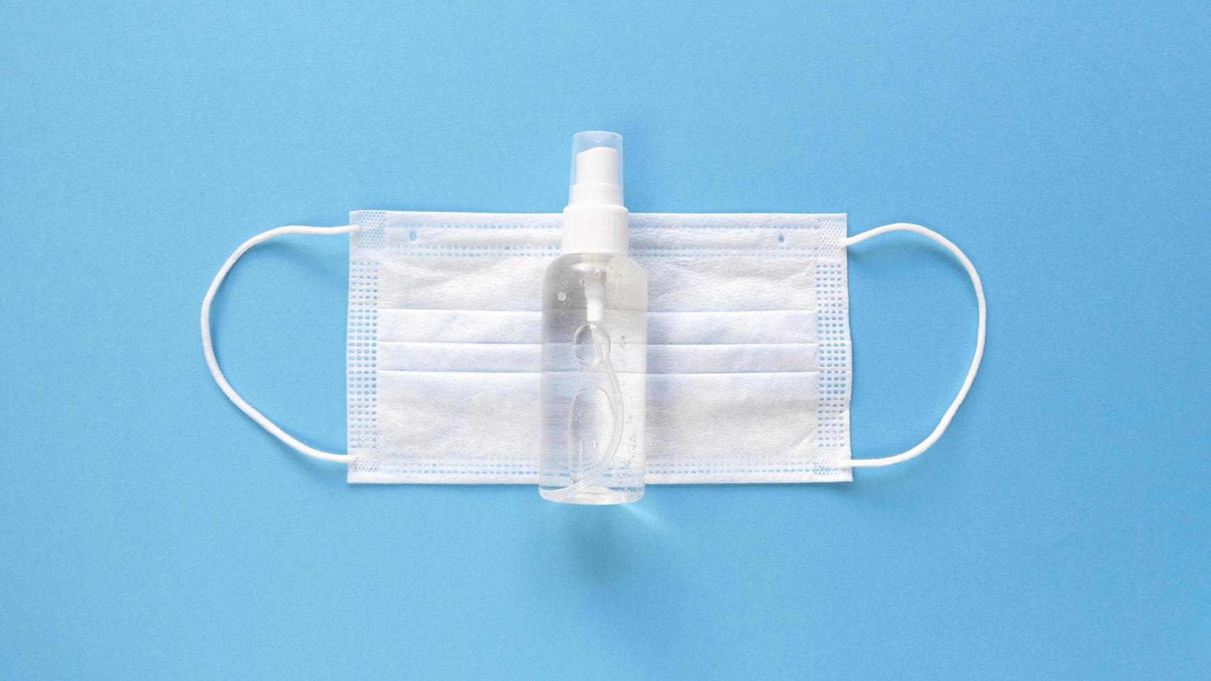 White medical mask and hand sanitizer in a transparent bottle with a spray cap at the middle of a blue background, simple flat lay with pastel paper texture medical concept photo