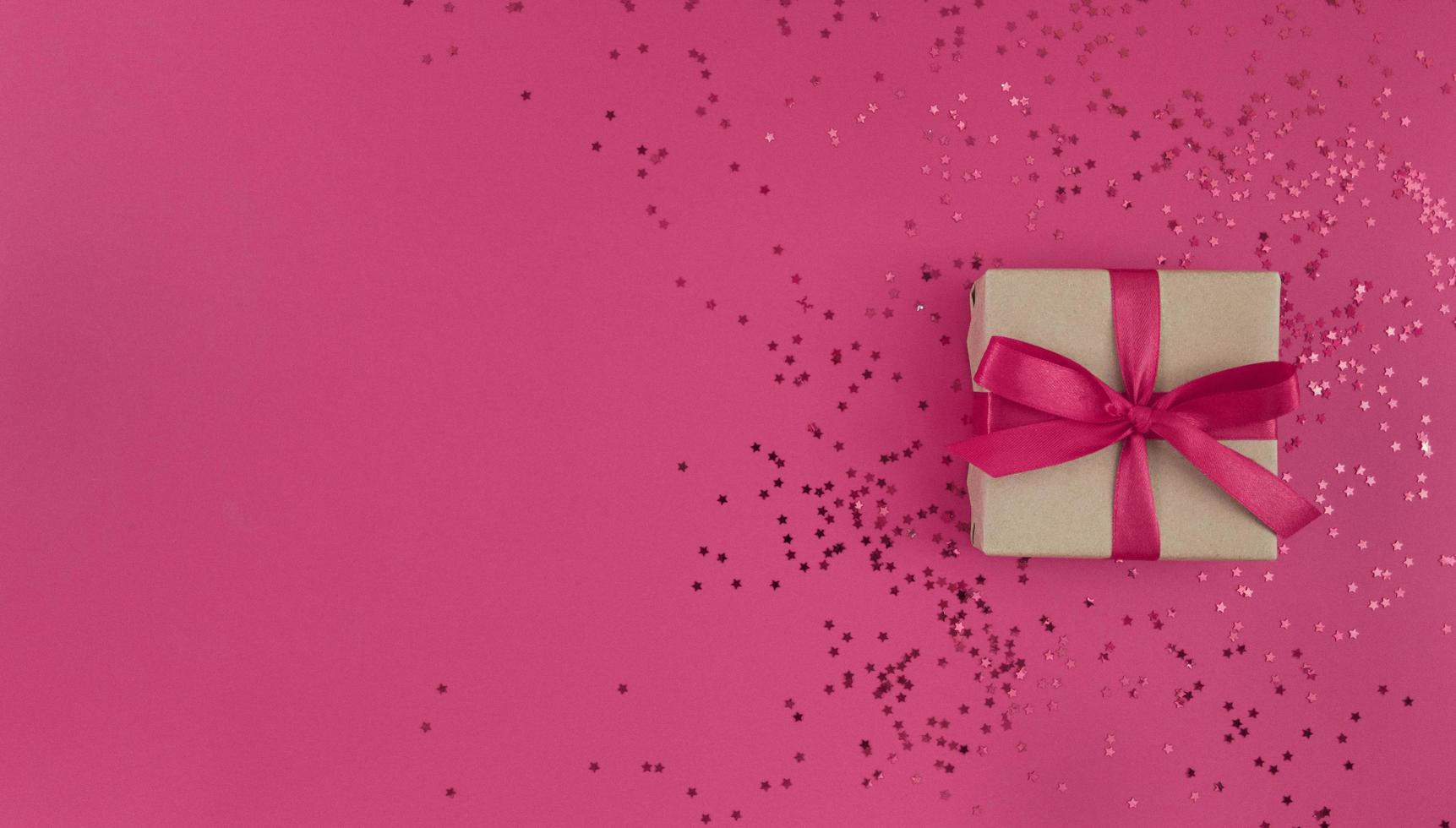 Gift box wrapped in craft paper with a pink ribbon bow and confetti on a pink background, monochrome festive flat lay with copy space photo