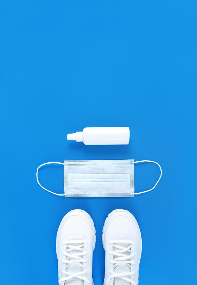 White medical mask, trainers, and hand sanitizer on a blue background, monochrome vertical flat lay photo