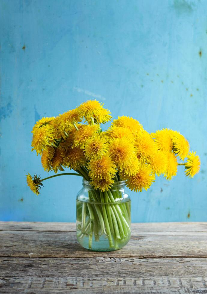 Bouquet of dandelions in glass jar on wooden table with light blue backdrop photo