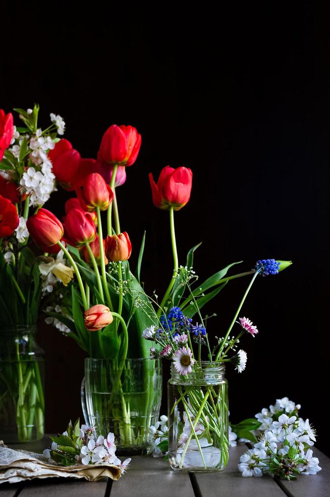 Naturaleza muerta con ramos de tulipanes rojos, margaritas de campo, muscaris en frascos de vidrio, flores de cerezo en la mesa de madera sobre fondo oscuro foto