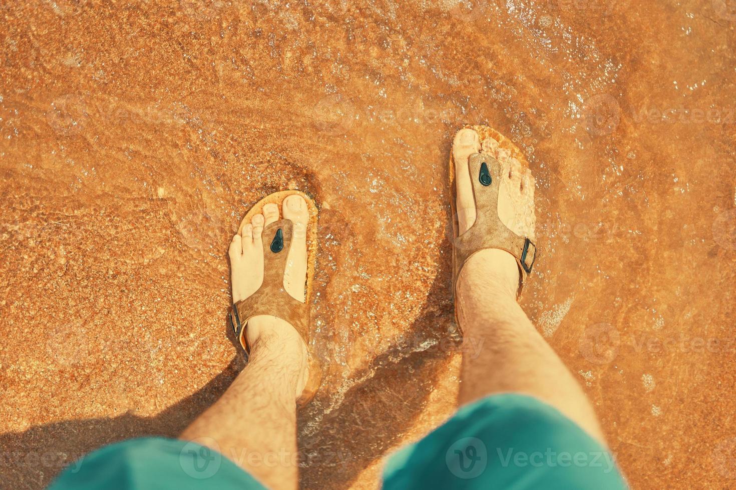 Persona en pantalones cortos de jeans se para y mira sus pies en la arena que se lava por el mar foto