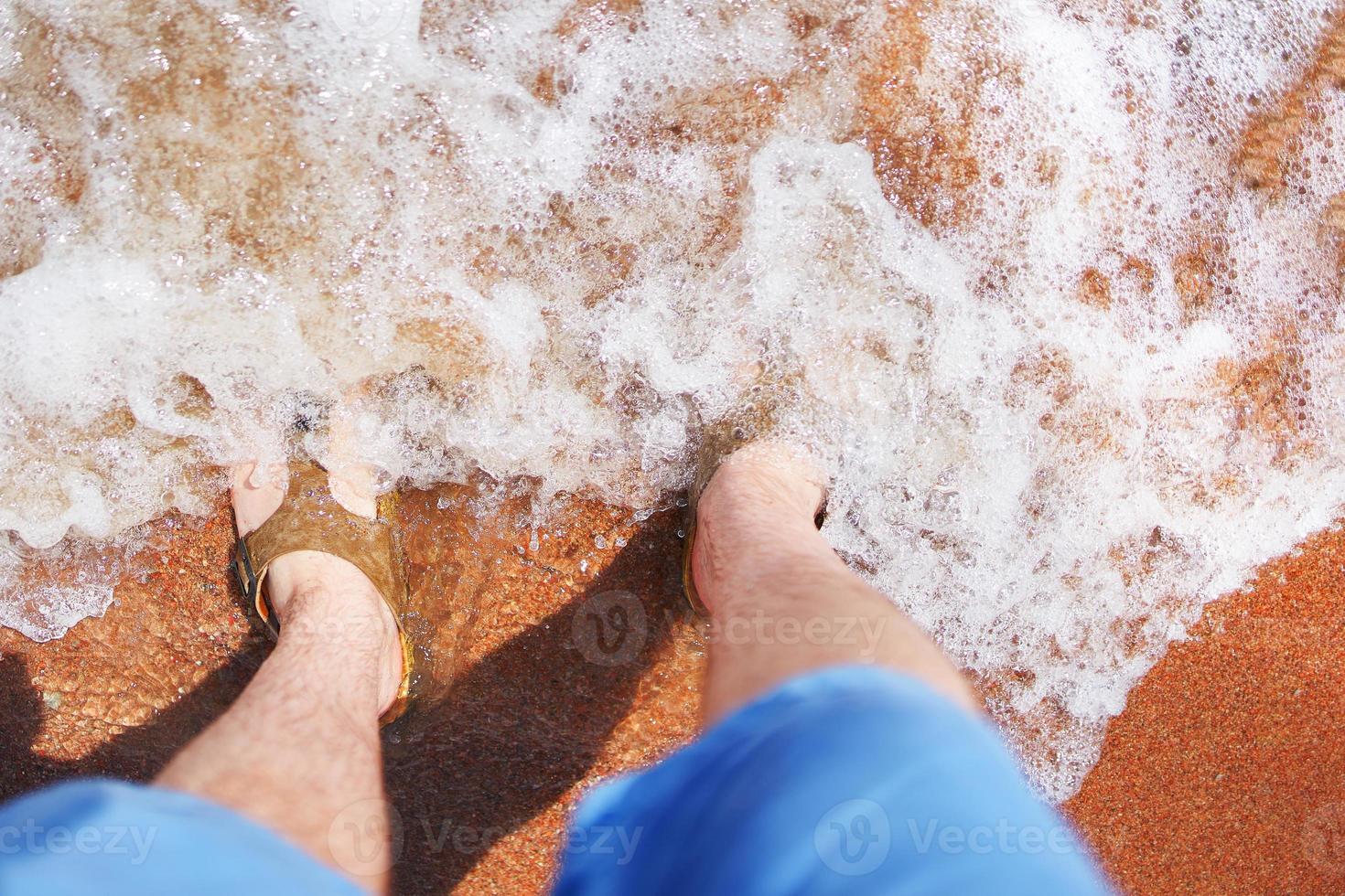Man in flip-flops stands in sand in an ocean wave photo