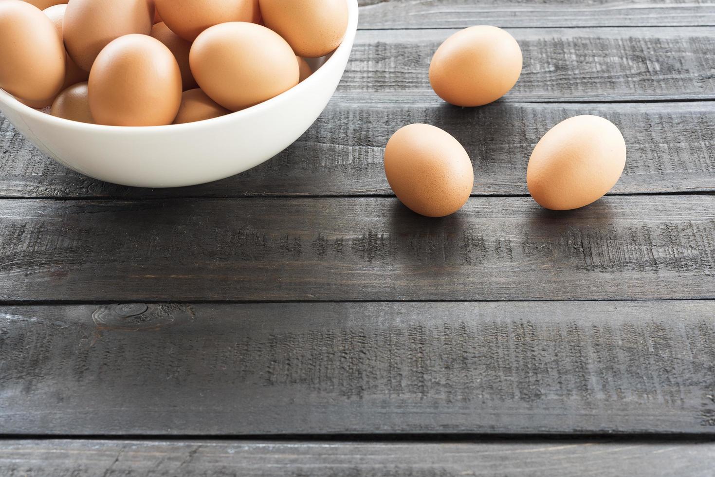 Huevos de gallina en un recipiente blanco y tres huevos de gallina afuera sobre una mesa de madera negra foto