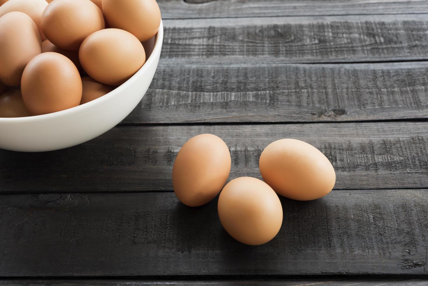 Huevos de gallina en un recipiente blanco y tres huevos de gallina afuera sobre una mesa de madera negra foto