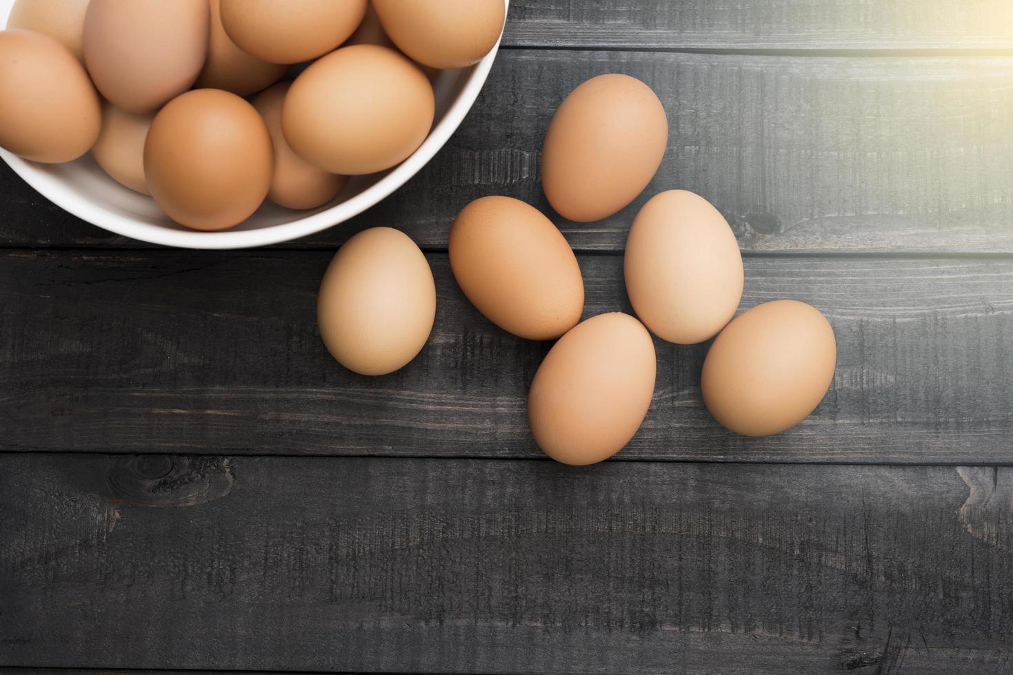 Huevos de gallina frescos en un recipiente blanco y seis huevos afuera sobre una mesa de madera negra foto