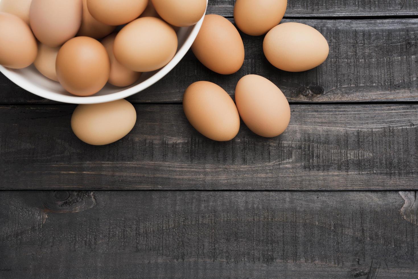 Huevos de gallina frescos en un recipiente blanco y seis huevos afuera sobre una mesa de madera negra foto