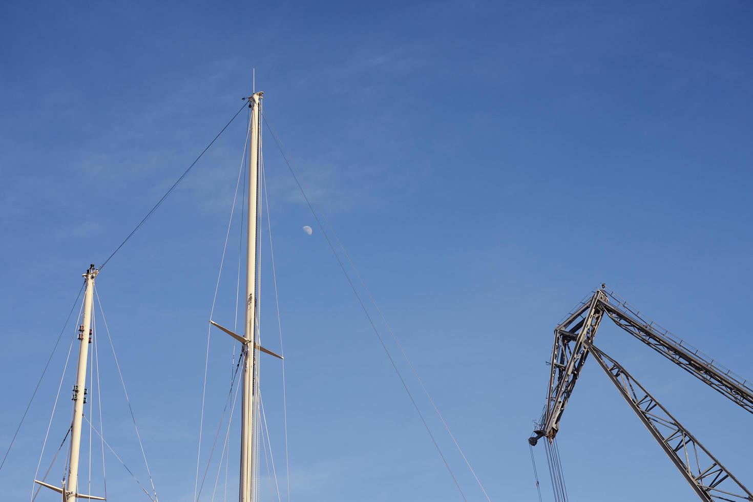Boat masts and a crane against a blue sky with the moon photo