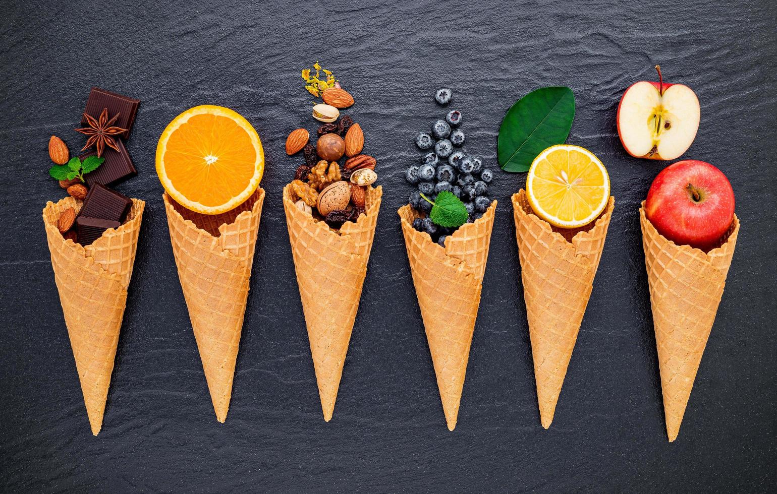 Various ingredients for ice cream flavors in cones showing blueberries, lime, pistachio, almonds, orange, chocolate, vanilla, and coffee set up on a dark stone background. Summer and sweet menu concept. photo