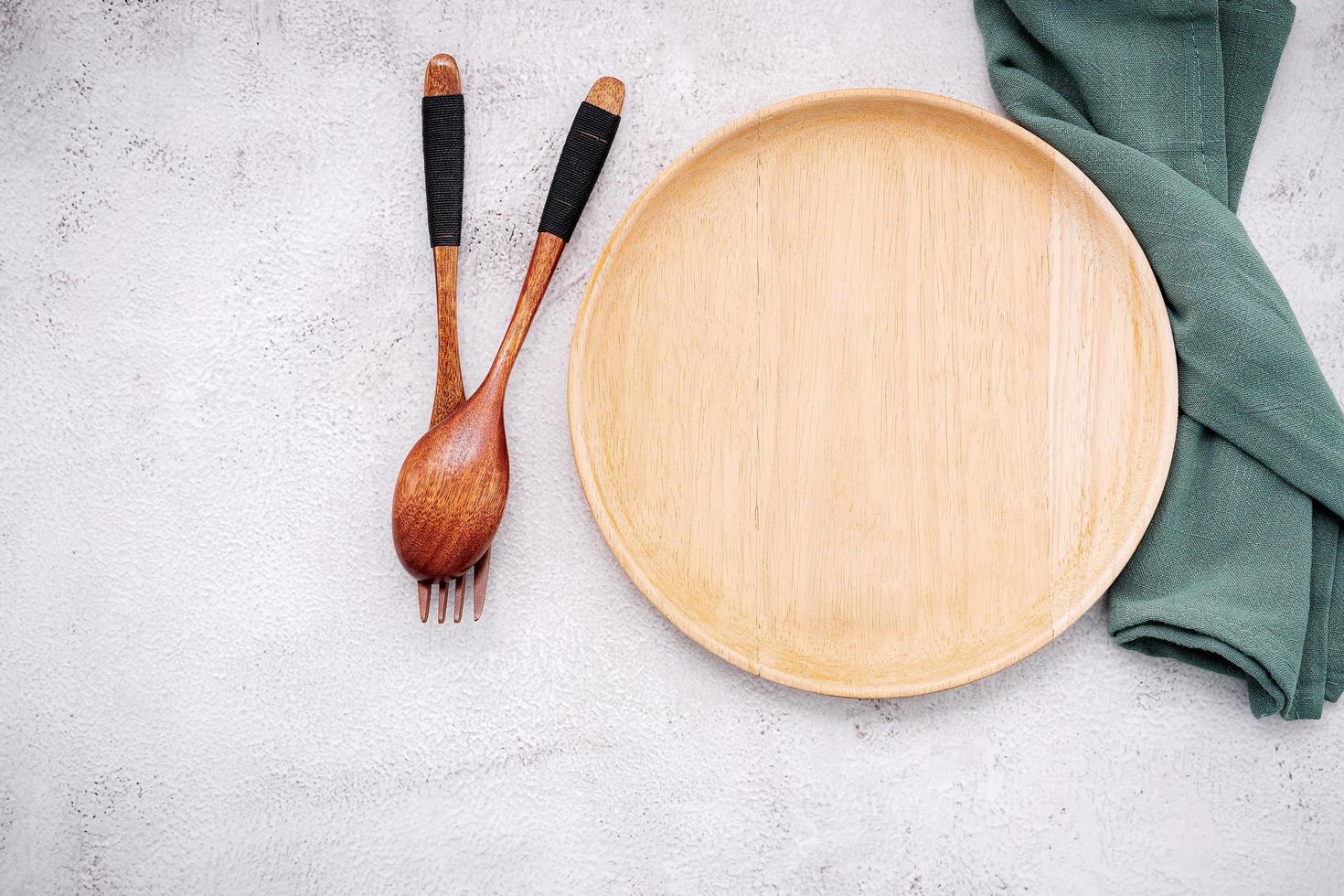 Empty wooden plate and a green linen napkin with a wooden spoon and fork on a white concrete background photo