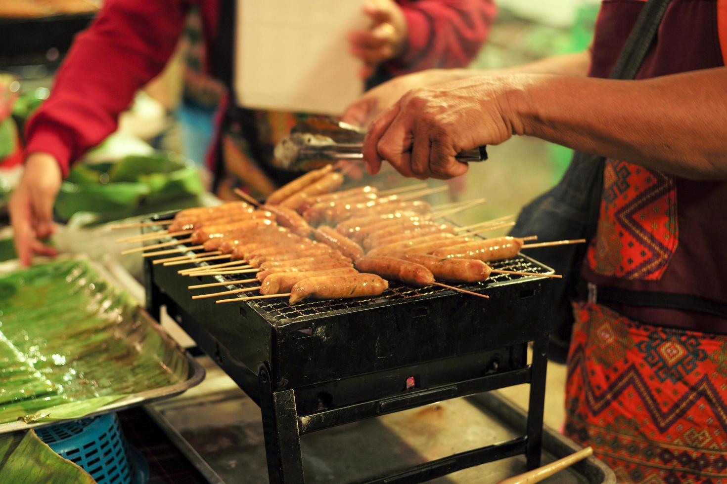 Closeup fila de salchichas de carne en la parrilla metálica con movimiento de manos borrosas y vendedor en segundo plano. foto