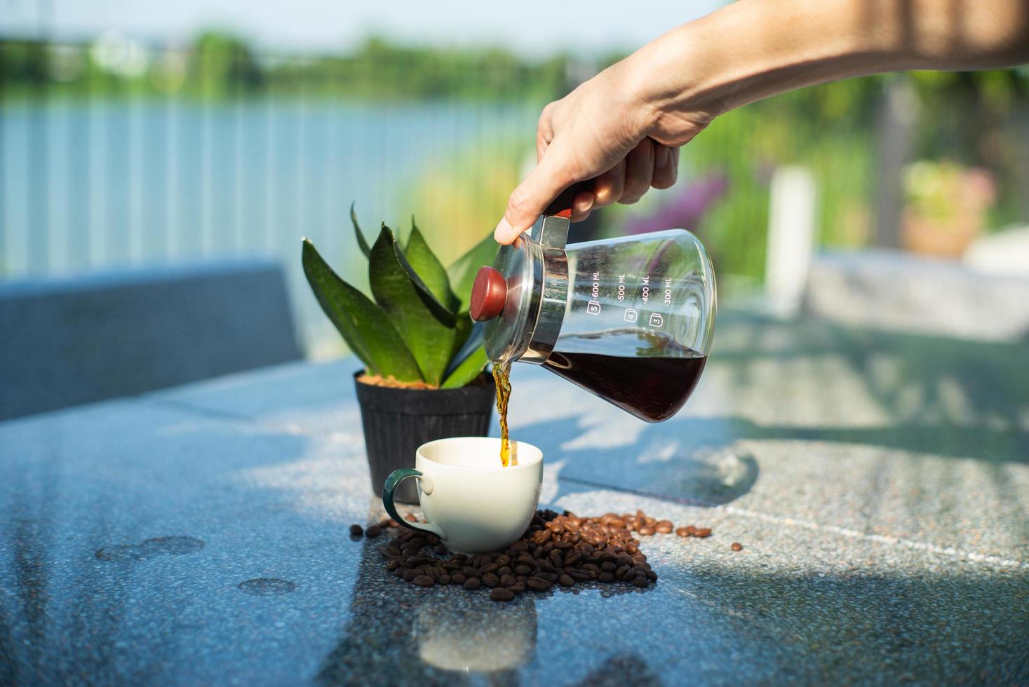 Hand of barista pouring the coffee from the glass jar into the cup at the outdoor garden photo