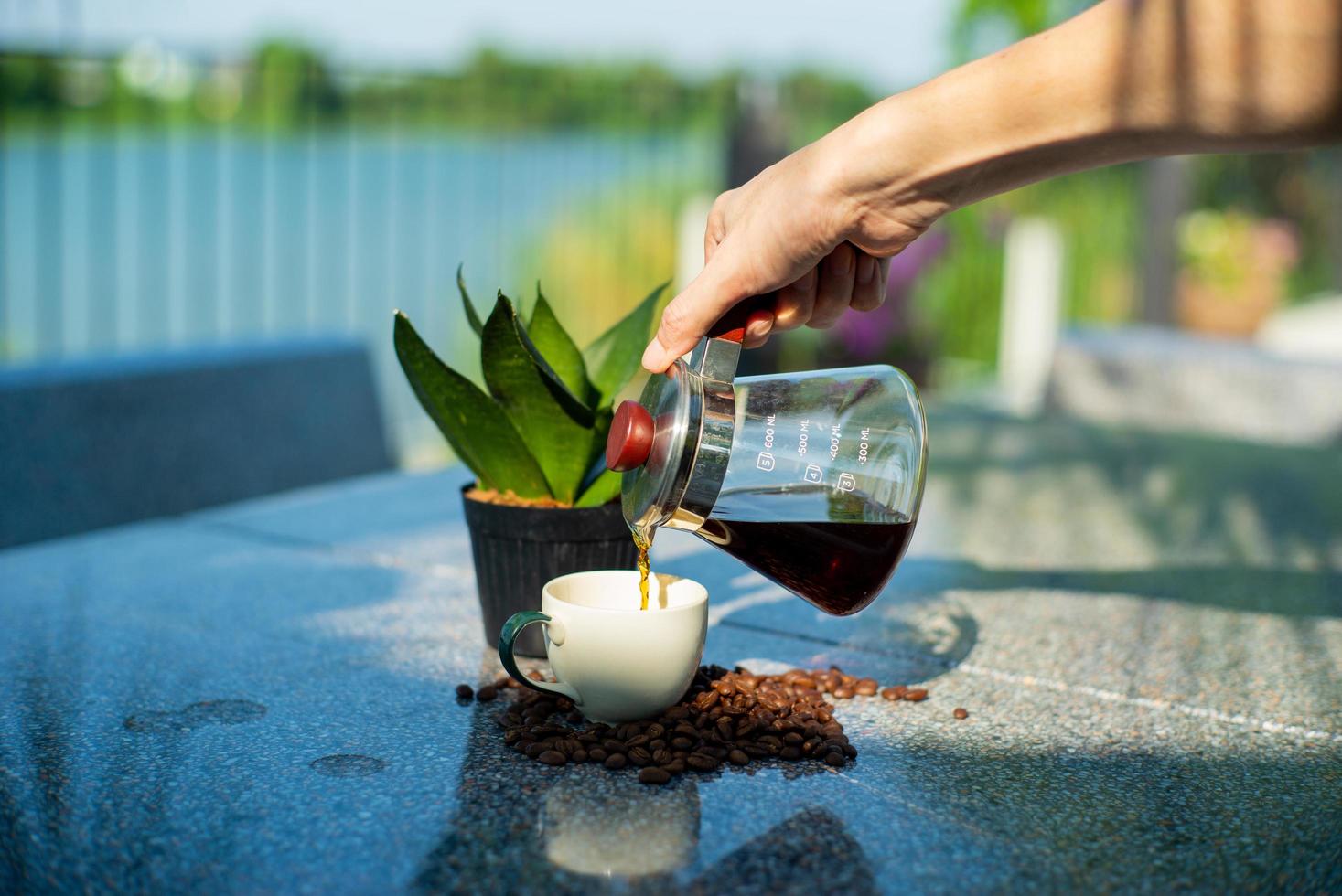 Hand of barista pouring the coffee from the glass jar into the cup at the outdoor garden photo