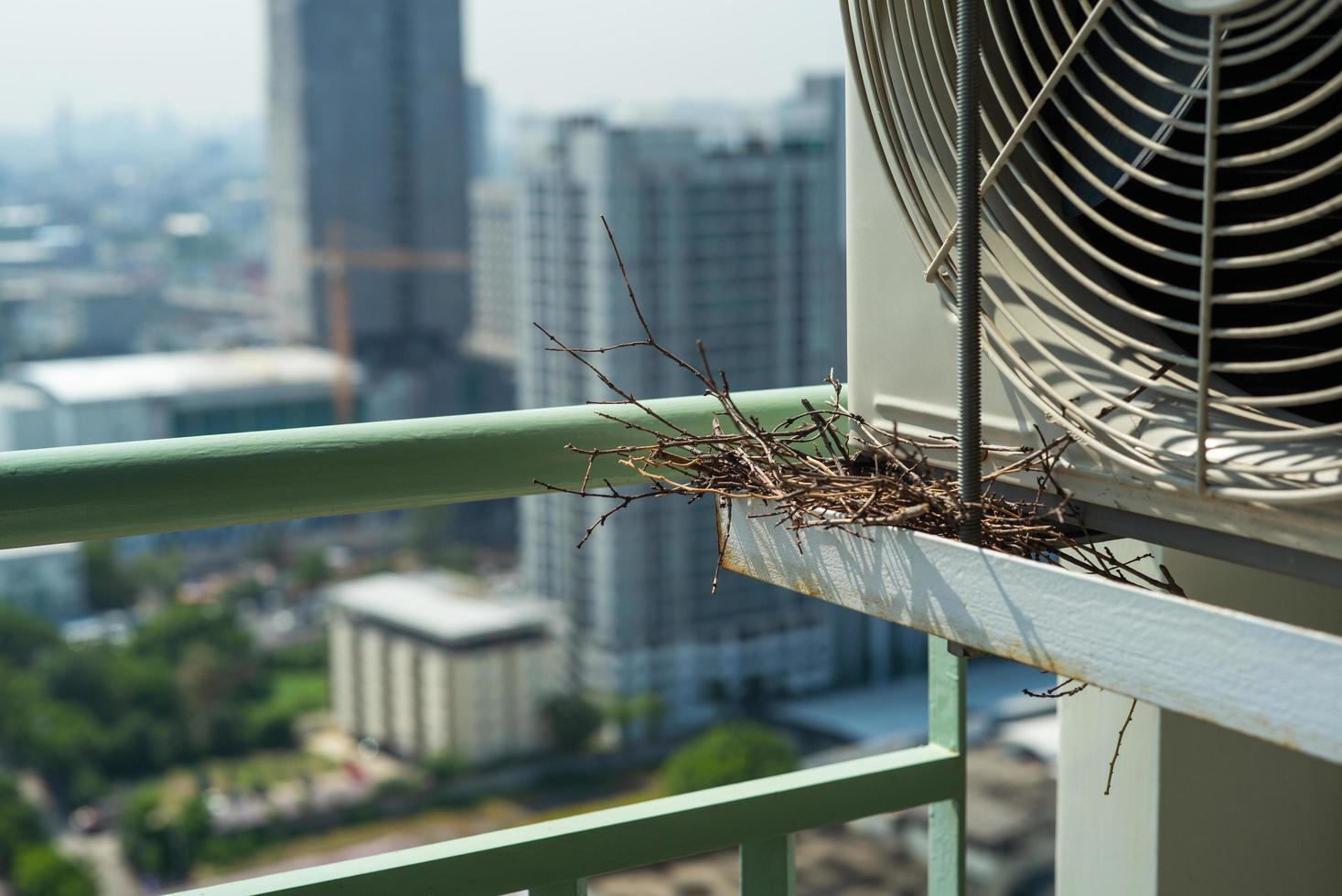 Closeup bird nest on the steel cage of air conditioner at the terrace of high condominium with blurred cityscape background in sunshine morning photo