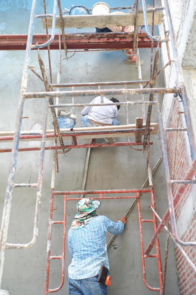 Grupo de trabajadores de pie sobre los andamios de acero y construye un muro de cemento enlucido en la casa en construcción foto