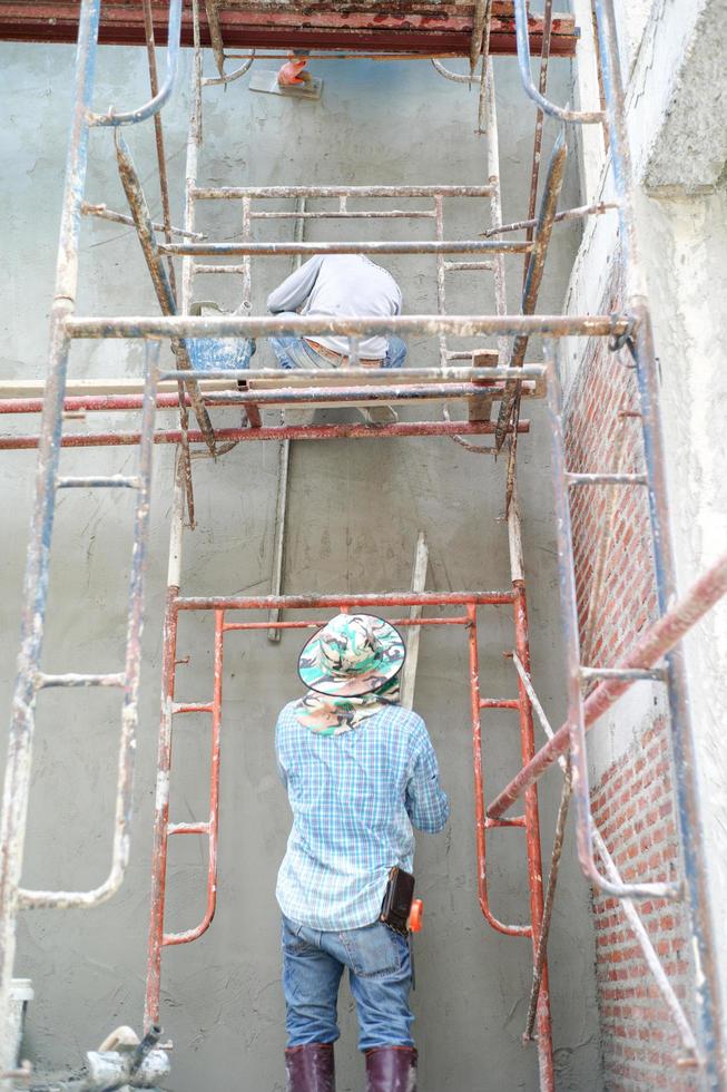 Group of workers stand on the steel scaffolding and builds plastered cement wall in the house under construction photo