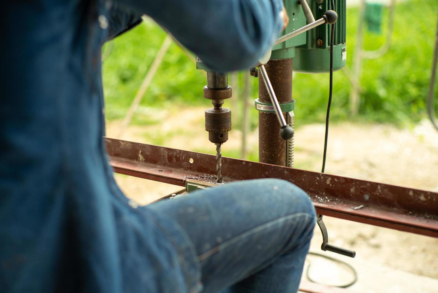 Primer enfoque selectivo en la espalda retrato de trabajador controla la máquina perforadora eléctrica para perforar el orificio en la barra de acero en ángulo en el sitio de construcción foto