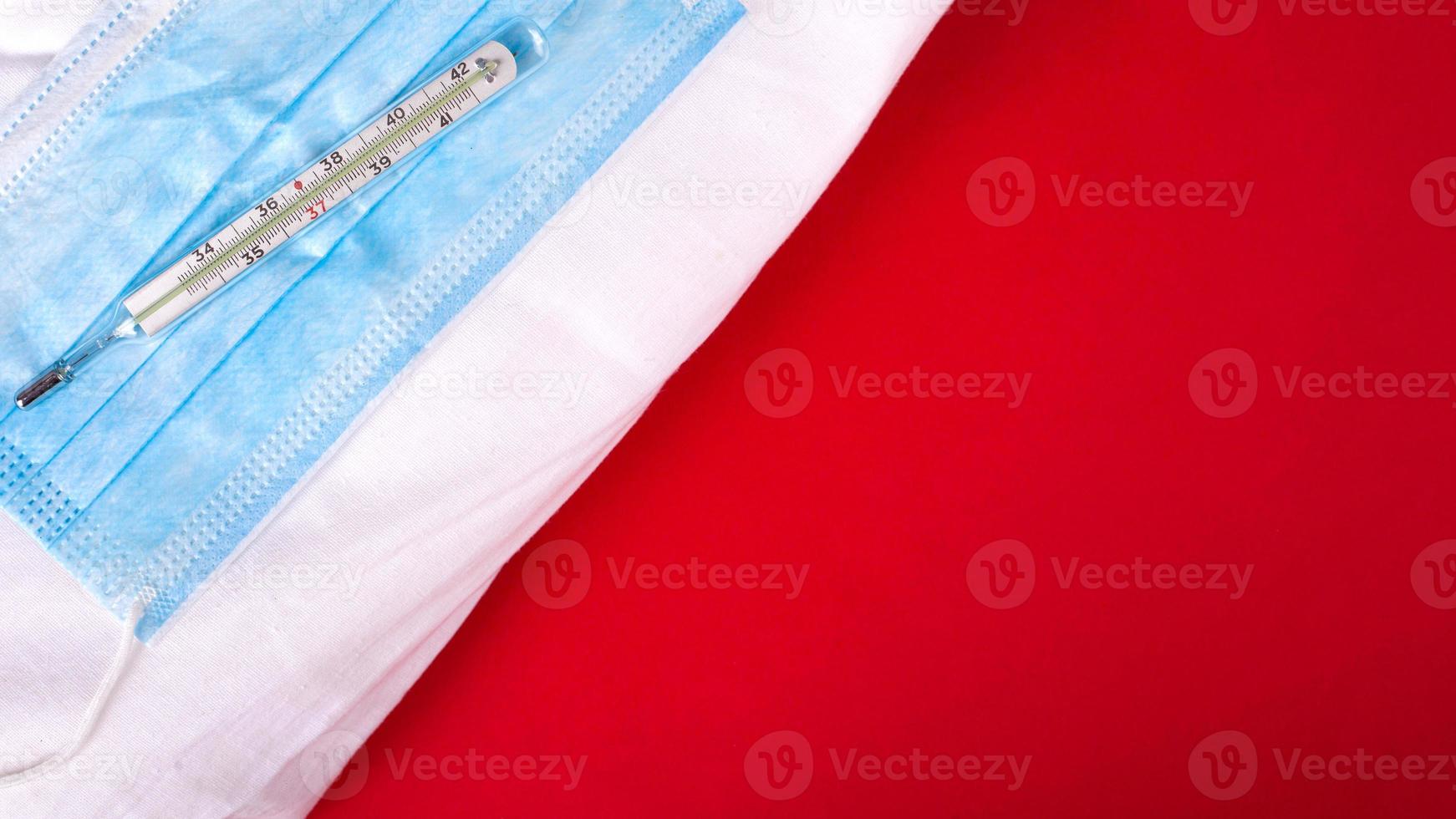 Máscara médica y termómetro en una bata de médico sobre un fondo rojo. foto