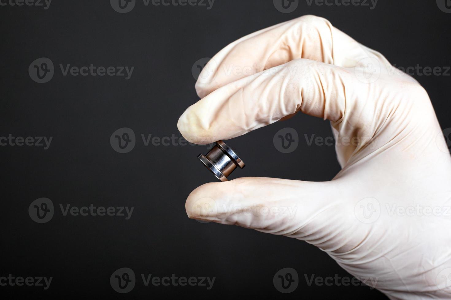 Hand in a white disposable glove holds a piercing tunnel on a dark background photo