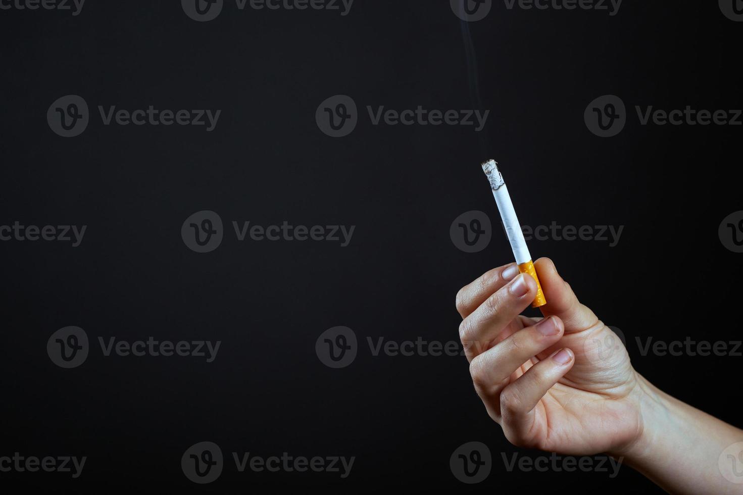 La mano sostiene un cigarrillo encendido sobre un fondo oscuro foto