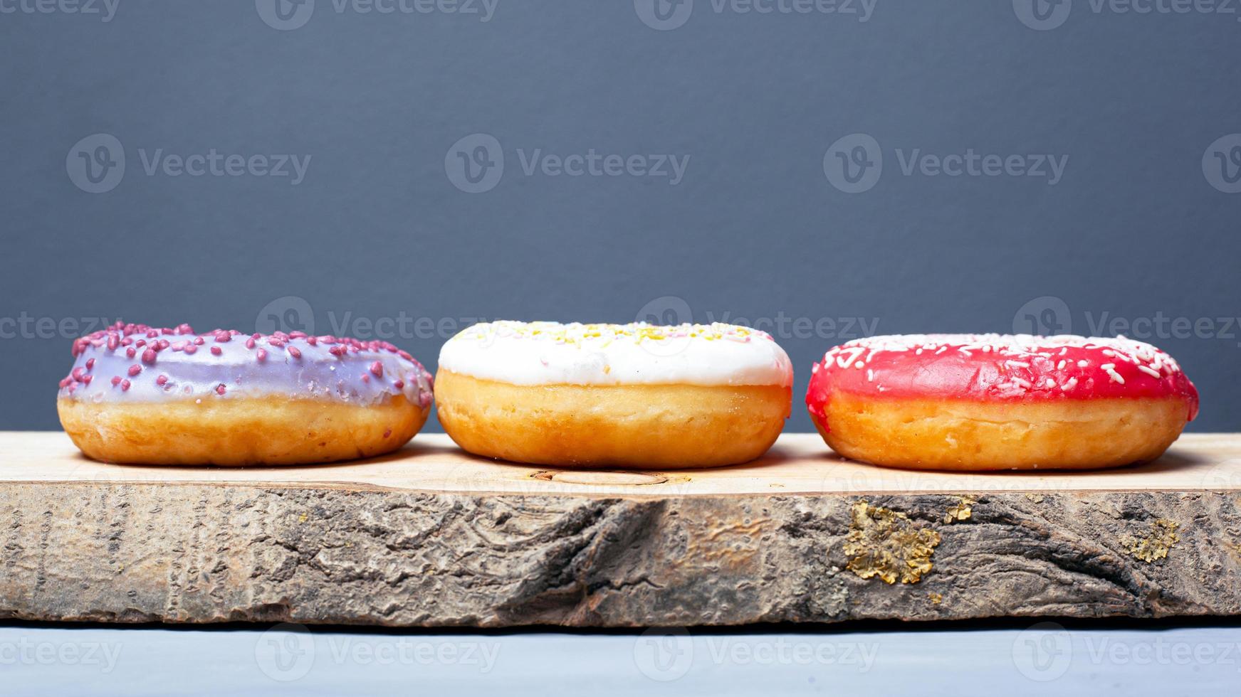 Three assorted glazed donuts of different colors on a wooden plank on a gray background photo