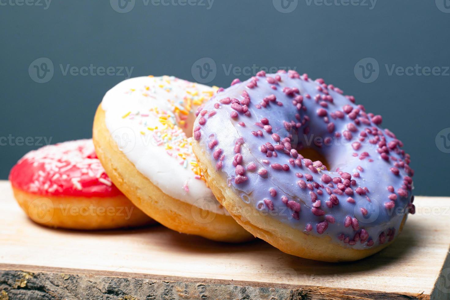 Donuts con glaseado rojo, blanco y morado multicolor sobre un soporte de madera sobre un fondo gris foto