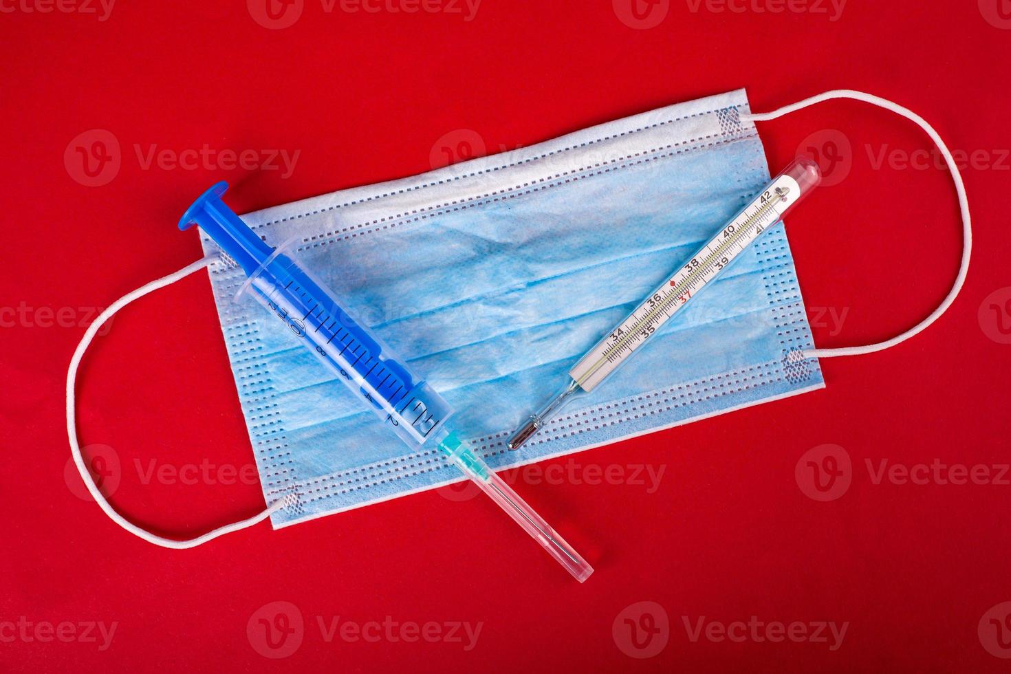 Mascarilla protectora, jeringa de vacuna y termómetro sobre fondo rojo. foto