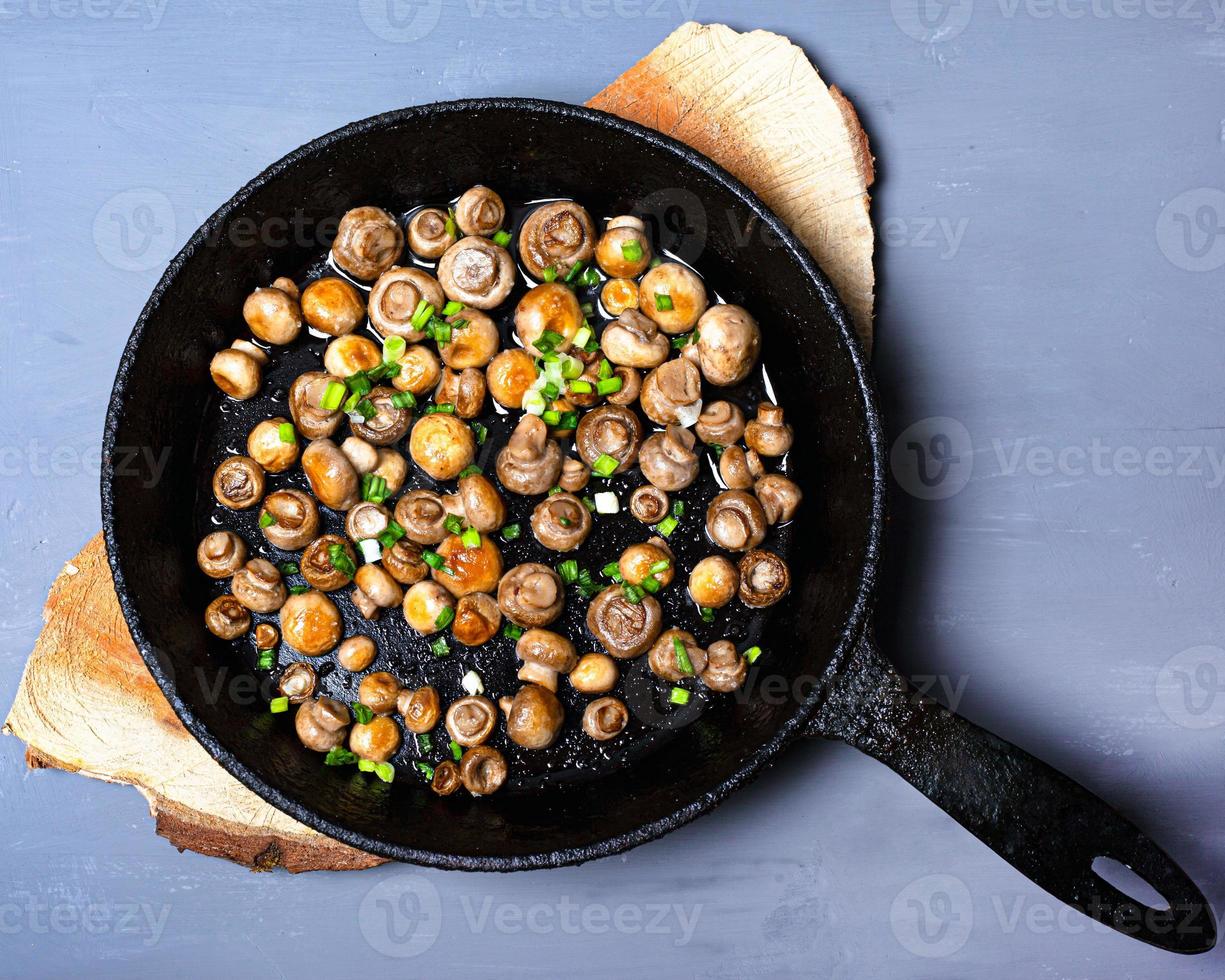 Fried champignon mushrooms in a cast-iron pan with fresh green onions on a wooden stand photo