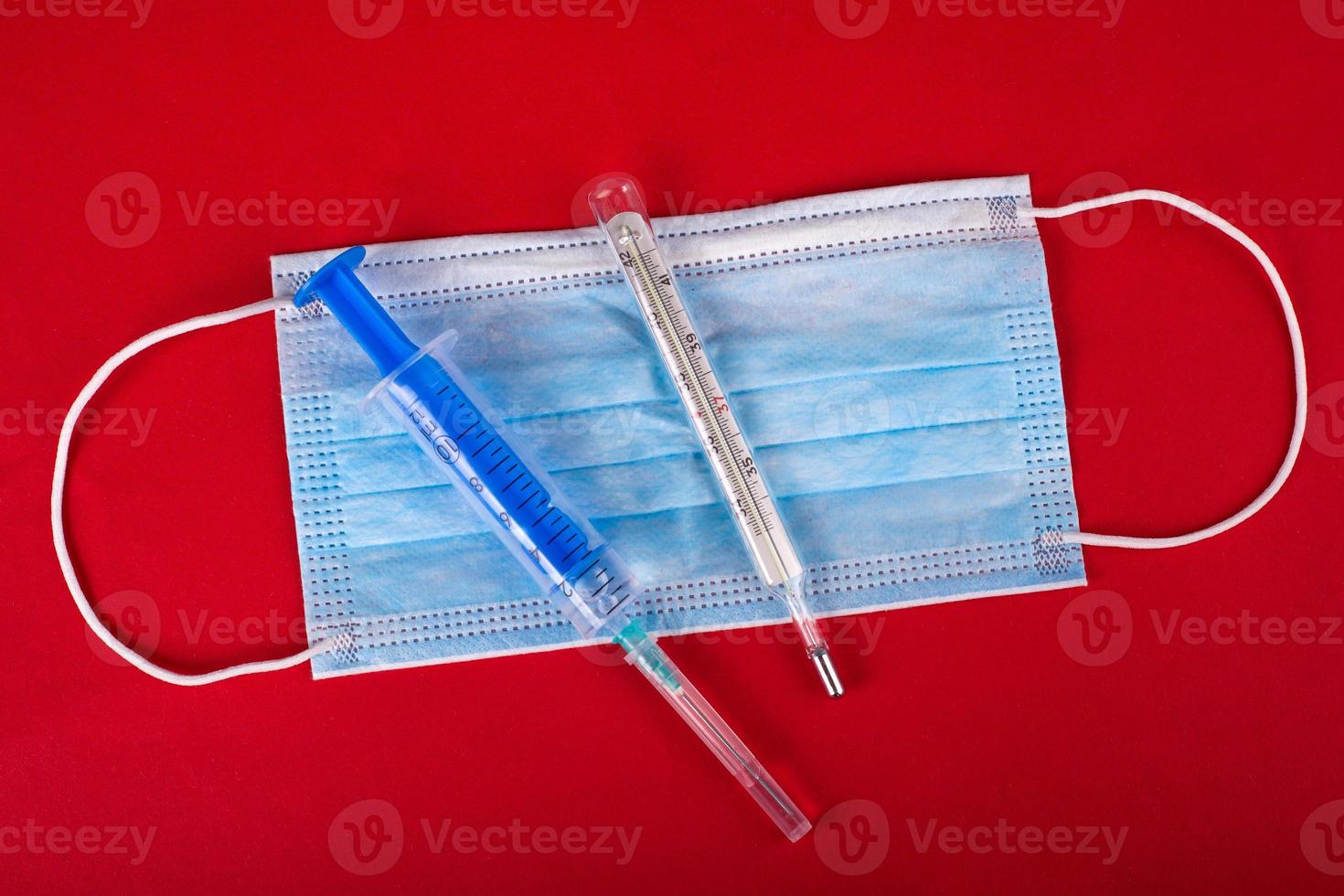 Máscara protectora médica, termómetro y jeringa con una vacuna sobre un fondo rojo. foto
