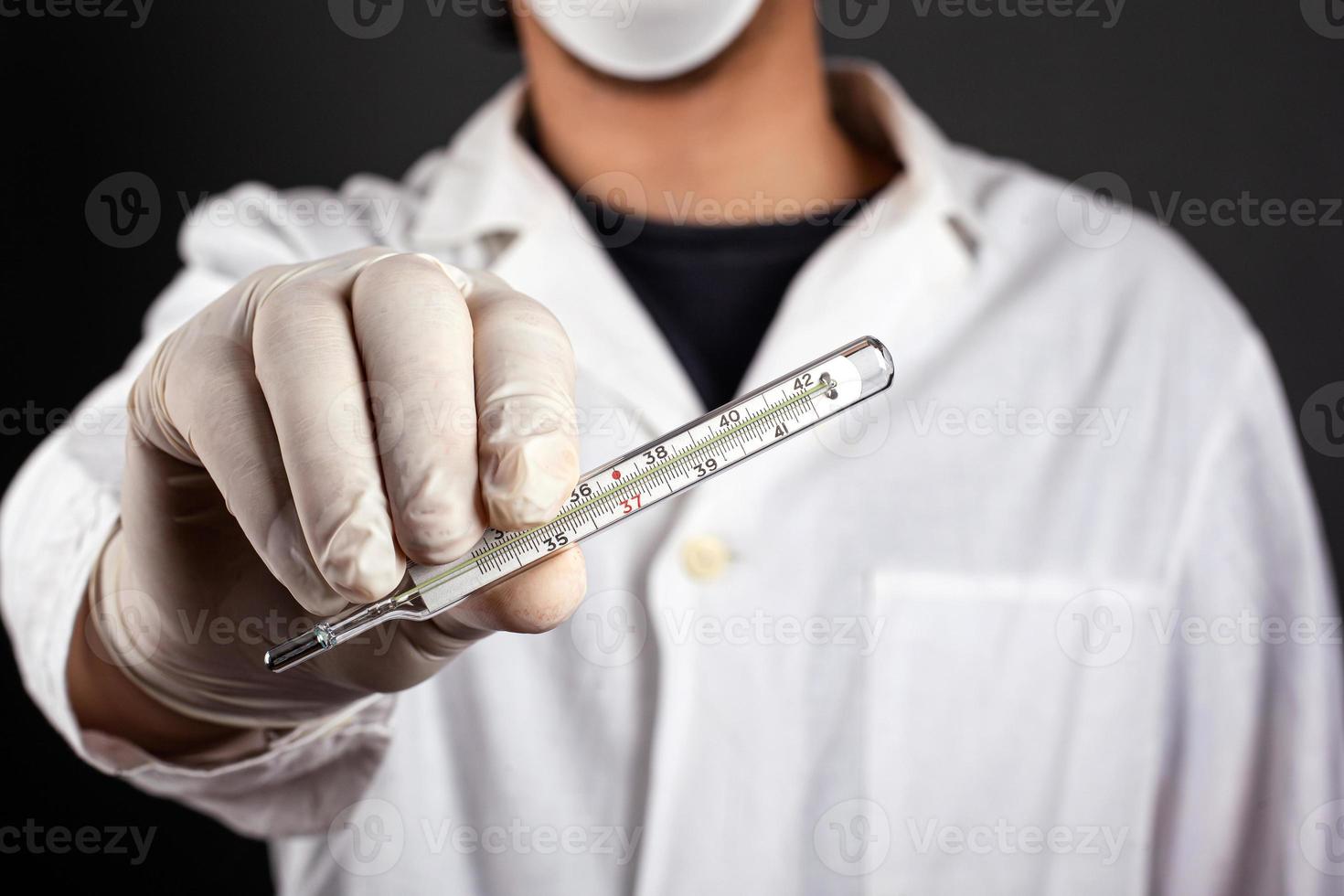 Doctor in a white coat in a mask and gloves holds in hand a thermometer photo