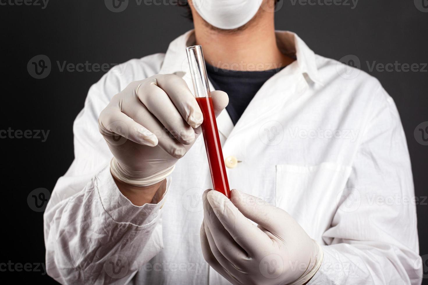 Doctor en bata blanca máscara médica y guantes estériles manos sosteniendo un tubo de ensayo con sangre infecciosa sobre un fondo oscuro foto