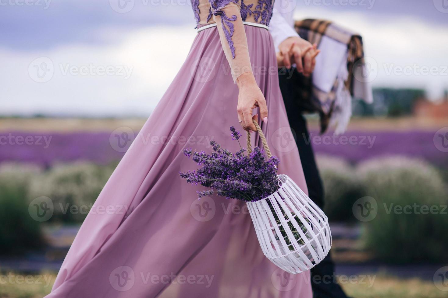 hermosa mujer joven con un vestido azul sostiene un ramo de flores de lavanda en una canasta mientras camina al aire libre a través de un campo de trigo al atardecer en verano. provenza, francia. imagen en tonos con espacio de copia foto