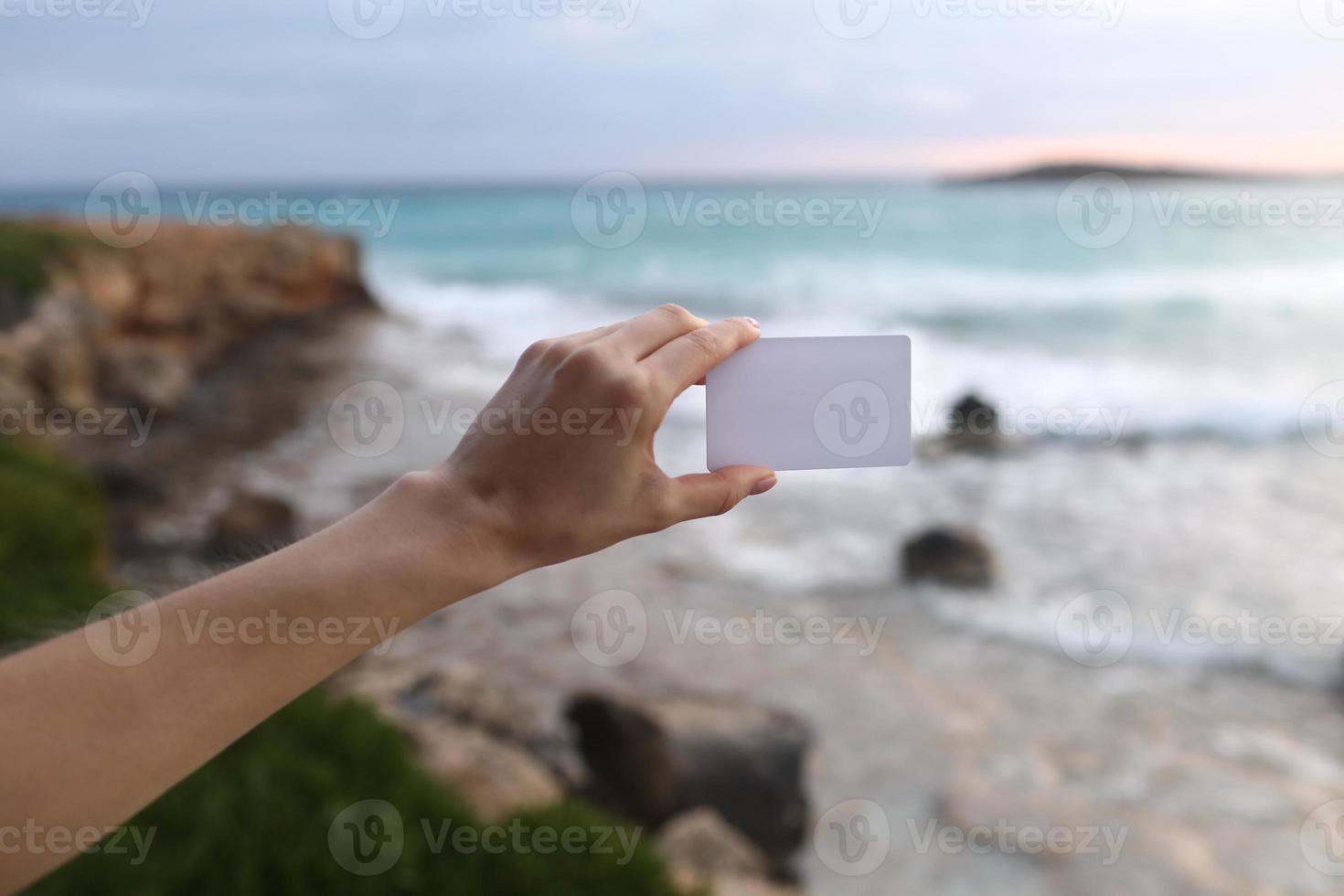 Female hand holding a white business card or sticky note with place for text on the beach background and the sea making waves. Selective focus photo