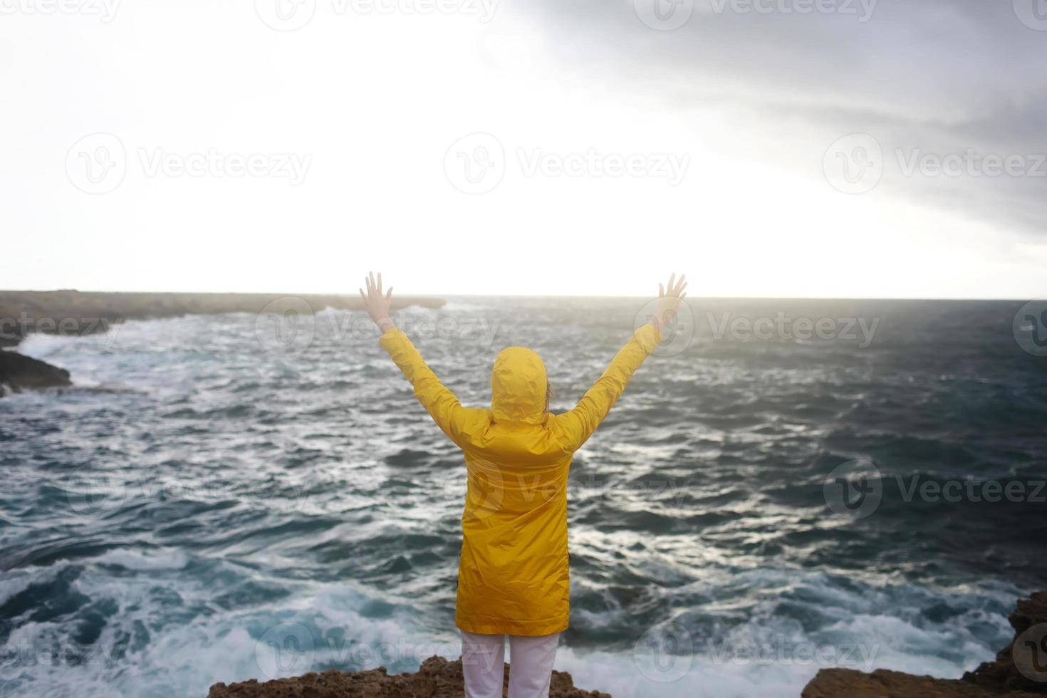 Mujer joven vestida con un impermeable amarillo de pie con los brazos extendidos mientras disfruta del hermoso paisaje marino en un día lluvioso en una playa rocosa en un clima primaveral nublado foto