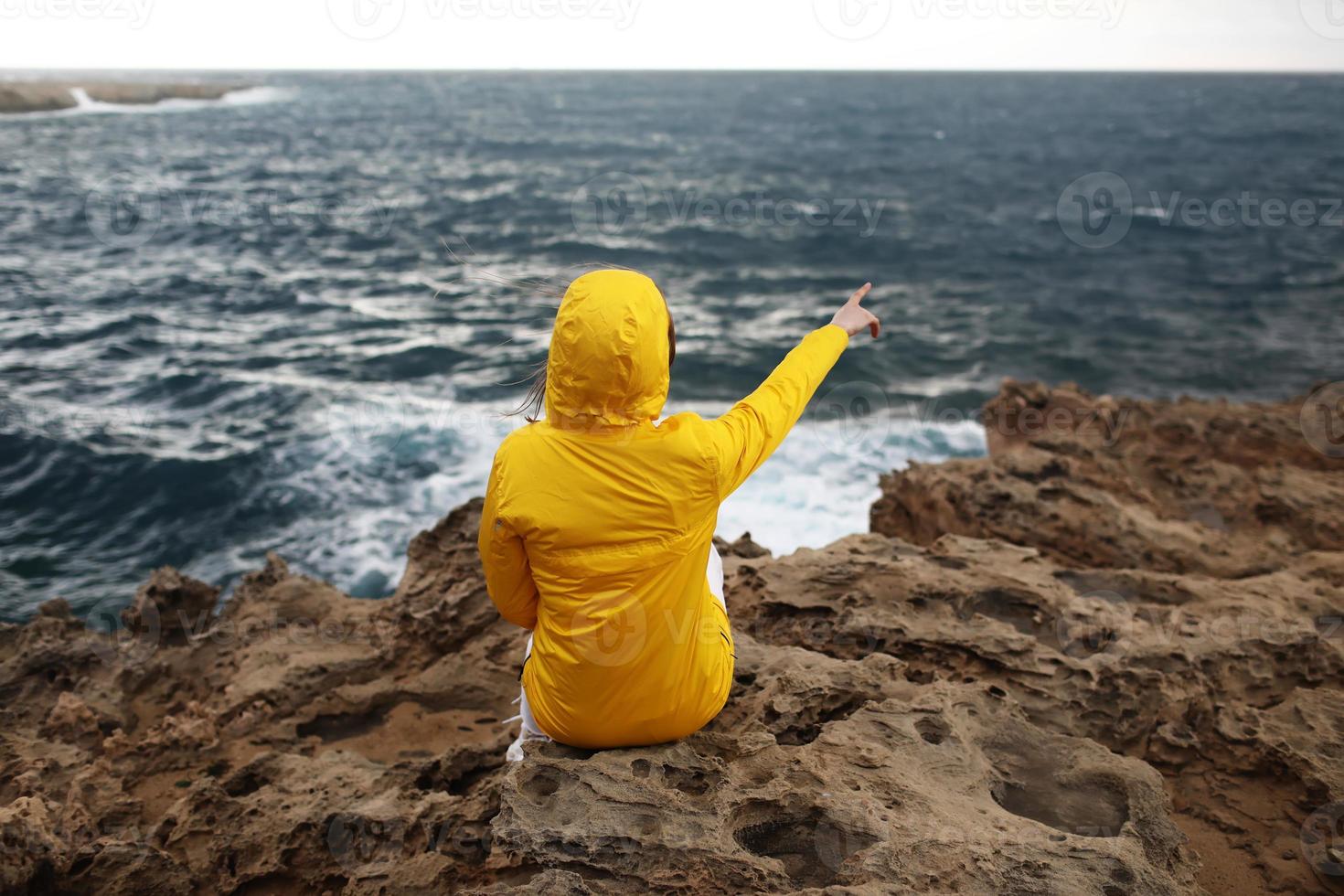 mujer joven con un impermeable amarillo está sentada en el acantilado mirando las grandes olas del mar mientras disfruta del hermoso paisaje marino en un día lluvioso en la playa de rocas en un clima primaveral nublado. foto