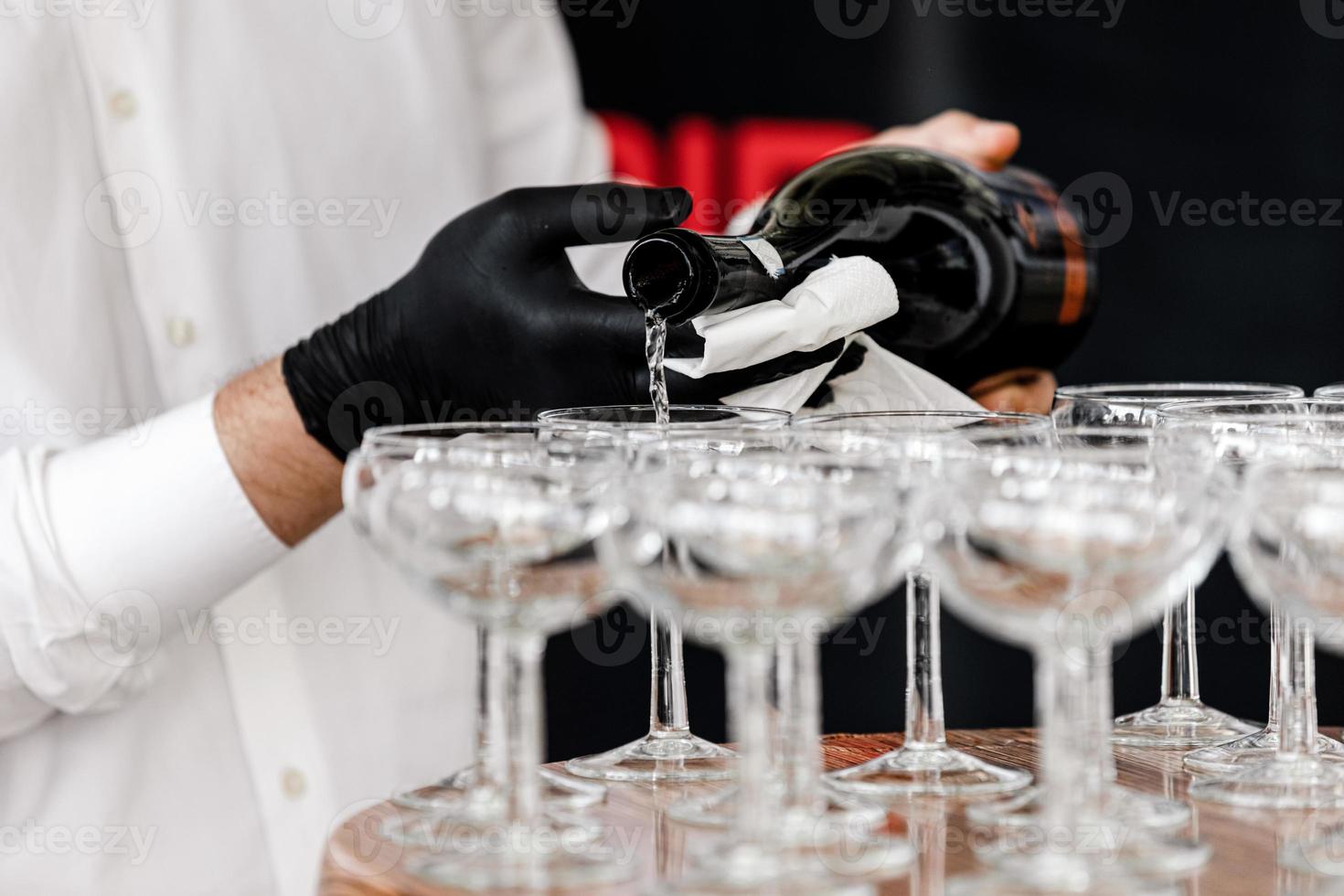 Hand with a black glove pouring champagne in the restaurant. Waiter in black gloves pouring champagne glasses on the wooden table. Selective focus. photo