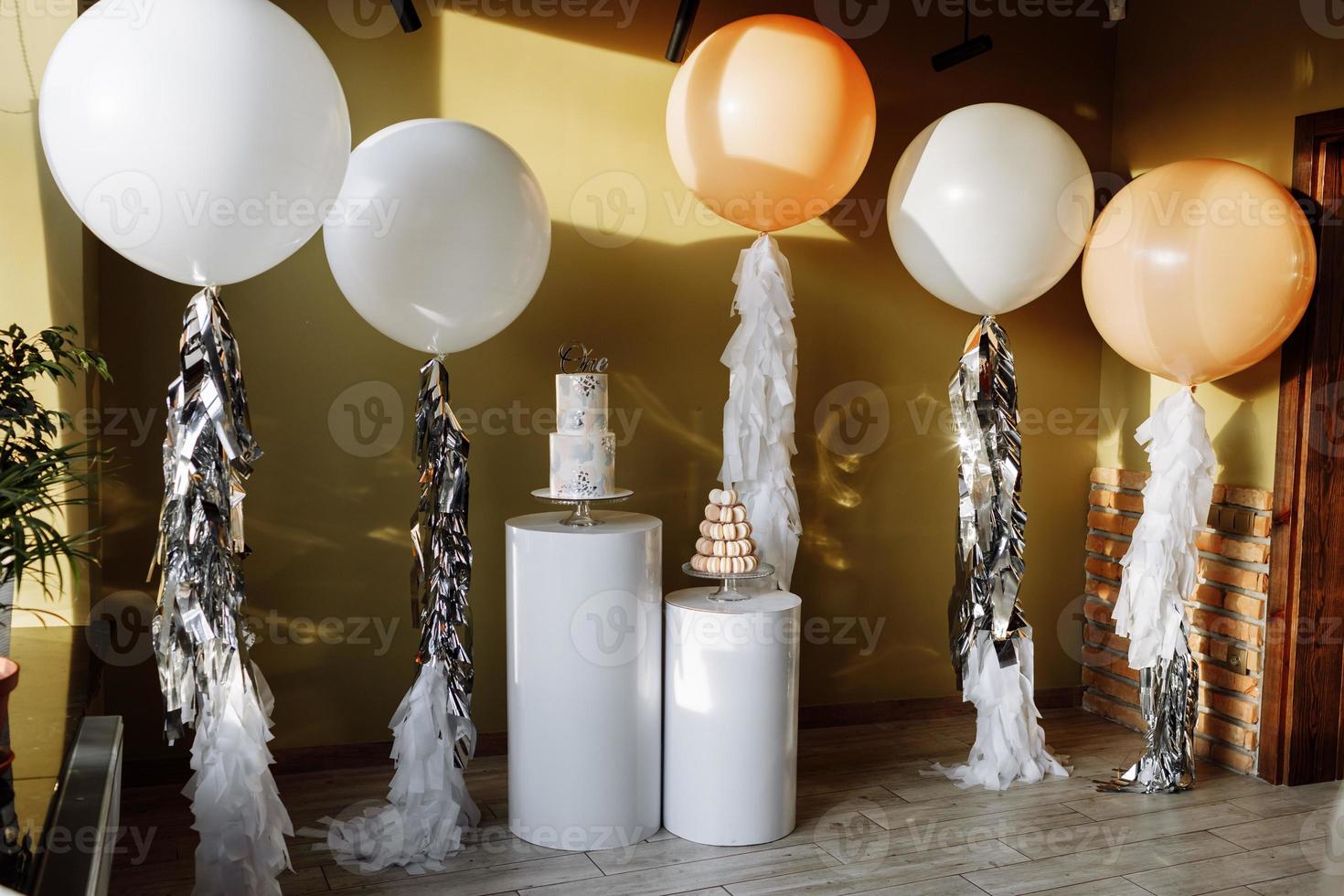 Decoration for birthday with a two-story cake and big white and beige balloons with tinsel. Candy bar with macaroon or macarons. Birthday party. Selective focus. photo