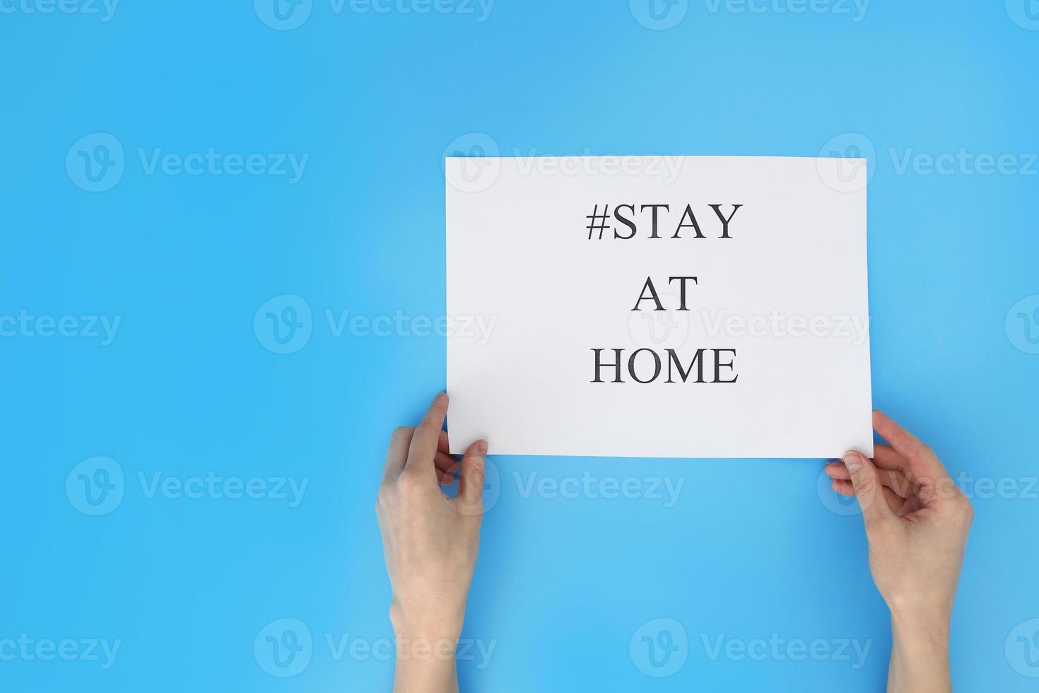 Stay at home concept. Woman hand holding a paper with text, hashtag stay at home, isolated on blue background. Coronavirus, COVID-19, self-quarantine, isolation. photo