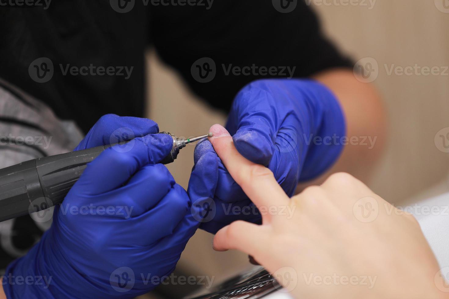 Close up shot of hardware manicure in a beauty salon. Manicurist in protective gloves is applying electric nail file drill to manicure on female fingers photo