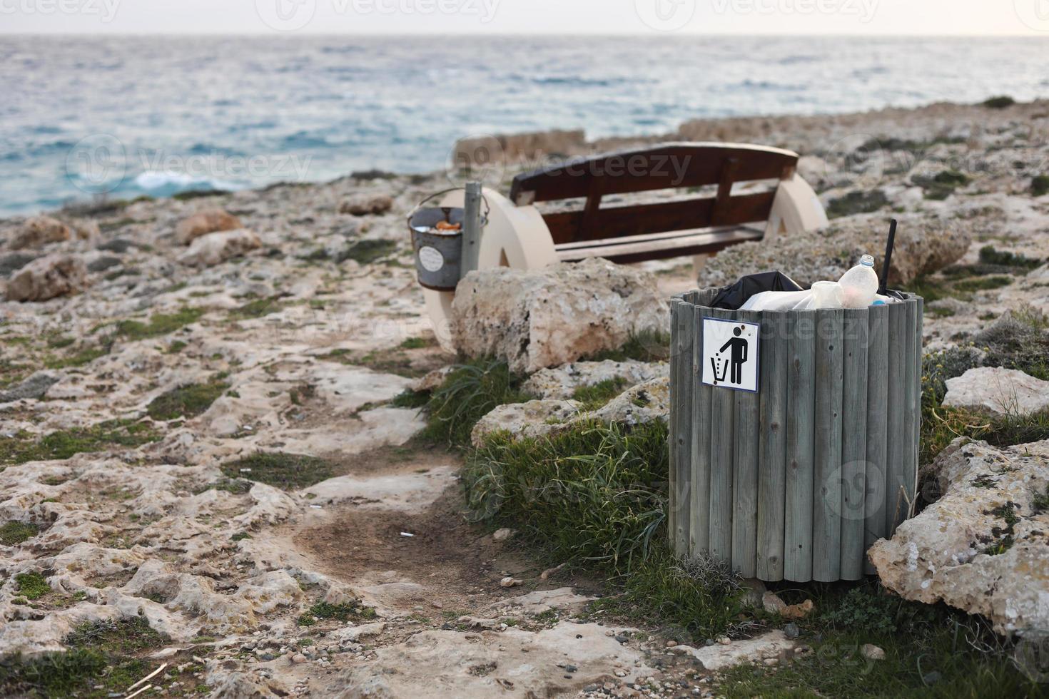 basura llena o papelera con botella de plástico, latas de cerveza y desechos orgánicos que muestran visiblemente la contaminación en las áreas costeras cercanas al mar. Banco de madera con balde con basura en la costa rocosa. foto